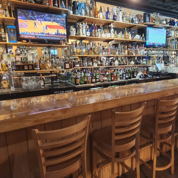 A well-stocked bar at Taxco Mexican Cuisine with numerous tequila bottles, bar stools, and TVs showing sports.