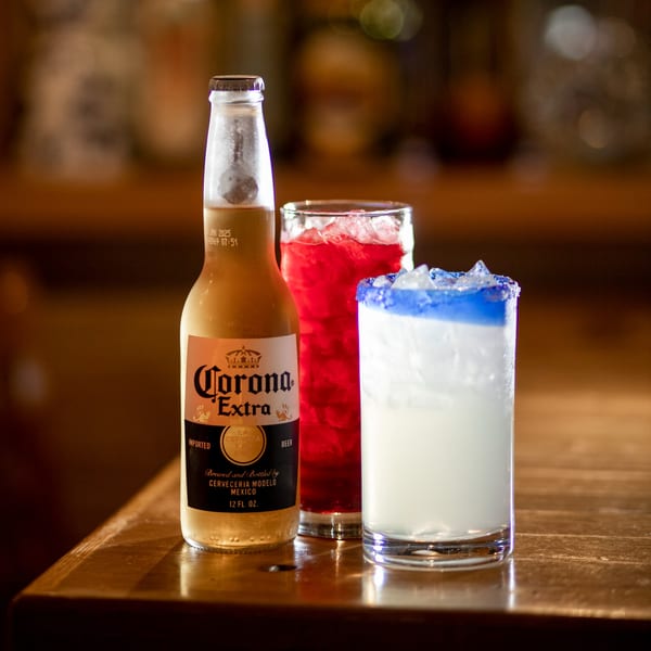 Corona Extra beer bottle, red drink, and blue-rimmed white drink on a wooden bar.