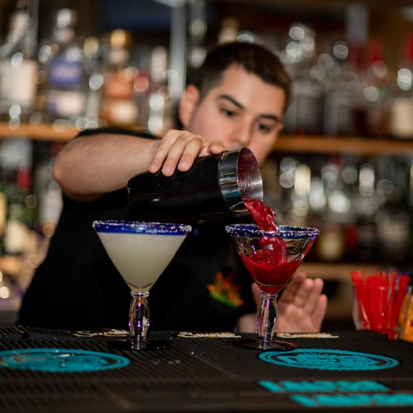 Bartender pouring a red margarita from a shaker into a glass, with another white margarita nearby.