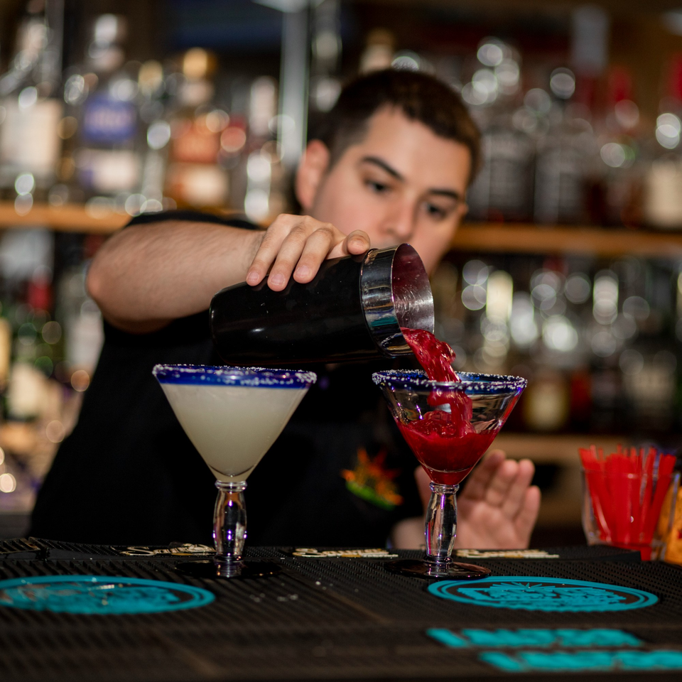 Bartender pouring a red margarita from a shaker into a glass, with another white margarita nearby.