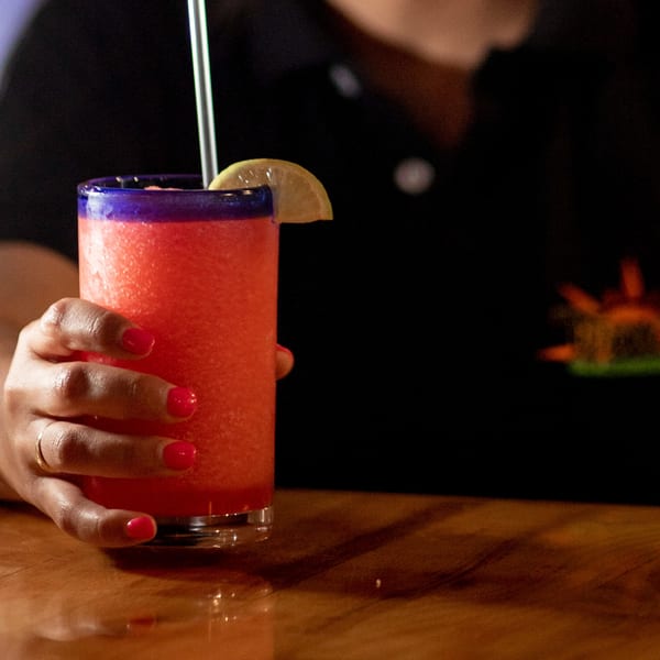 A hand with red nail polish holds a vibrant red frozen drink with a lime wedge and straw.