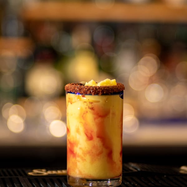 A vibrant yellow and red mango margarita with a chili-lime rim and pineapple chunks, on a bar.