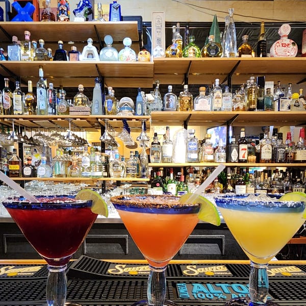 Three colorful margaritas with lime wedges and salt rims on a bar, with shelves of tequila bottles behind.